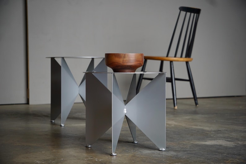 Puede incluir: Dos mesas auxiliares modernas y geom&eacute;tricas con acabado plateado. Cada mesa tiene un dise&ntilde;o angular &uacute;nico y una parte superior circular. Un cuenco de madera descansa sobre una mesa. Una silla negra con asiento de madera est&aacute; al fondo.