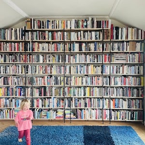 May include: A large, custom-built bookshelf filled with books of various sizes and colors. The shelves are supported by black metal frames and extend to the ceiling. A young child in pink clothing is walking in front of the bookshelf. A blue rug is on the floor.