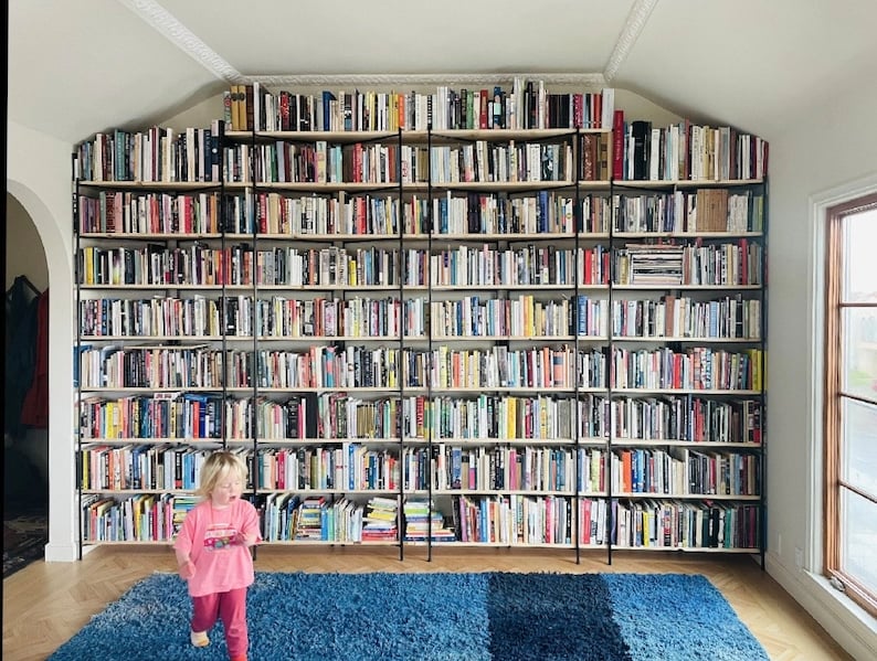 May include: A large, custom-built bookshelf filled with books of various sizes and colors. The shelves are supported by black metal frames and extend to the ceiling. A young child in pink clothing is walking in front of the bookshelf. A blue rug is on the floor.