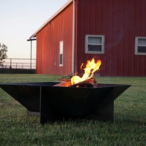 May include: A black, geometric fire pit with a burning fire, set on a grassy lawn. The flames are bright orange and yellow. A red barn is in the background.