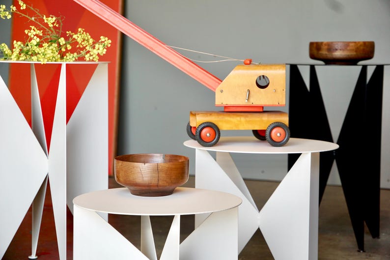 Puede incluir: Una gr&uacute;a de juguete de madera con ruedas rojas y una pluma roja se encuentra sobre una mesa blanca. En la imagen tambi&eacute;n hay otras mesas con dise&ntilde;os geom&eacute;tricos y cuencos de madera. El fondo presenta formas geom&eacute;tricas rojas y blancas.