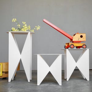 Puede incluir: Tres mesas auxiliares geom&eacute;tricas blancas con base en forma de X. Una mesa tiene flores amarillas, otra un juguete de gr&uacute;a de madera y una tercera est&aacute; vac&iacute;a. Un cami&oacute;n de juguete amarillo y una silla negra tambi&eacute;n est&aacute;n en la escena.