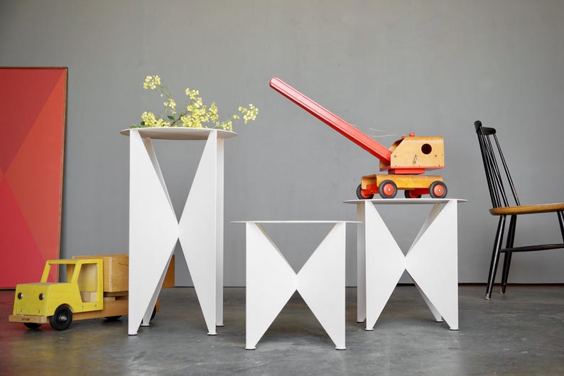 Puede incluir: Tres mesas auxiliares geom&eacute;tricas blancas con base en forma de X. Una mesa tiene flores amarillas, otra un juguete de gr&uacute;a de madera y una tercera est&aacute; vac&iacute;a. Un cami&oacute;n de juguete amarillo y una silla negra tambi&eacute;n est&aacute;n en la escena.