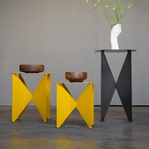 Puede incluir: Una escena interior moderna con tres mesas auxiliares. Dos son de color amarillo brillante con un dise&ntilde;o geom&eacute;trico, y una es negra. Cuencos de madera se encuentran sobre las mesas amarillas. Una silla negra y un jarr&oacute;n blanco con forma de mano y flores completan la decoraci&oacute;n.