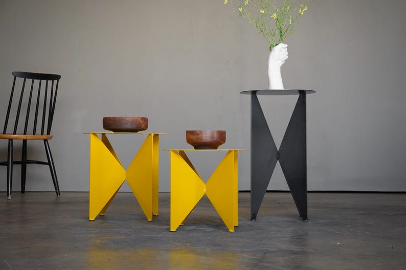 Puede incluir: Una escena interior moderna con tres mesas auxiliares. Dos son de color amarillo brillante con un dise&ntilde;o geom&eacute;trico, y una es negra. Cuencos de madera se encuentran sobre las mesas amarillas. Una silla negra y un jarr&oacute;n blanco con forma de mano y flores completan la decoraci&oacute;n.