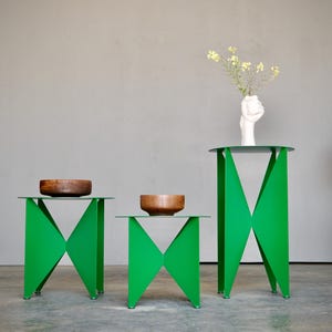 Puede incluir: Tres mesas auxiliares verdes modernas con dise&ntilde;os geom&eacute;tricos. Cada mesa tiene una tapa de cristal transparente y un cuenco de madera. Un jarr&oacute;n blanco con flores se encuentra sobre la mesa m&aacute;s alta. Una silla decorativa est&aacute; en el fondo.