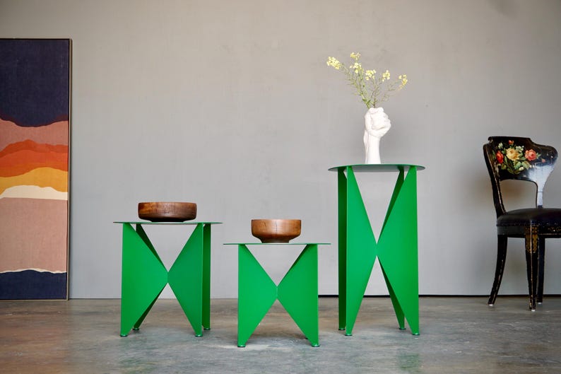 Puede incluir: Tres mesas auxiliares verdes modernas con dise&ntilde;os geom&eacute;tricos. Cada mesa tiene una tapa de cristal transparente y un cuenco de madera. Un jarr&oacute;n blanco con flores se encuentra sobre la mesa m&aacute;s alta. Una silla decorativa est&aacute; en el fondo.