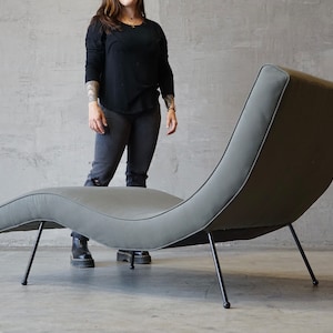 May include: A modern, curved chaise lounge in a muted gray fabric. The chair has a sleek design with thin, black metal legs. The overall aesthetic is minimalist and contemporary, suitable for a living room or lounge area.