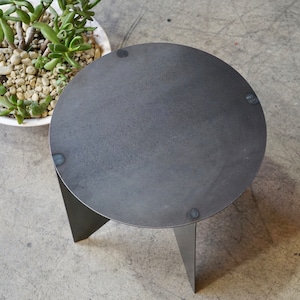 Puede incluir: Una peque&ntilde;a mesa auxiliar redonda de metal gris oscuro con tres patas anguladas. La mesa tiene una superficie texturizada y se muestra desde una perspectiva superior. Una maceta de cer&aacute;mica blanca con una planta suculenta est&aacute; en el fondo.