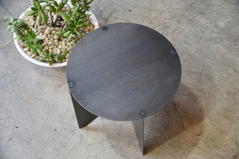 Puede incluir: Una peque&ntilde;a mesa auxiliar redonda de metal gris oscuro con tres patas anguladas. La mesa tiene una superficie texturizada y se muestra desde una perspectiva superior. Una maceta de cer&aacute;mica blanca con una planta suculenta est&aacute; en el fondo.