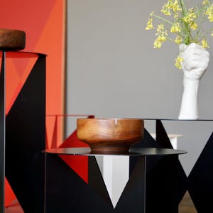 Puede incluir: Mesas auxiliares geom&eacute;tricas de metal negro con cuencos de madera. Un jarr&oacute;n blanco en forma de mano contiene flores amarillas. El fondo presenta un dise&ntilde;o geom&eacute;trico rojo y naranja.
