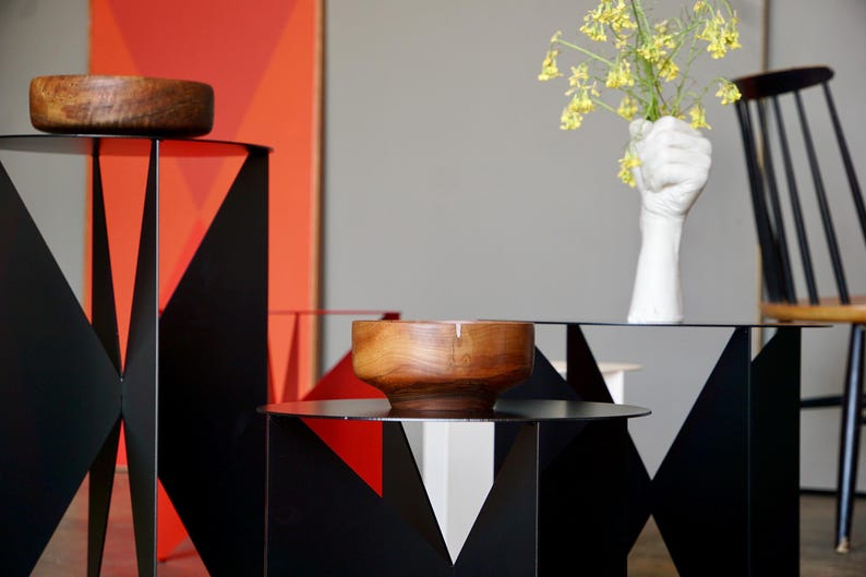 Puede incluir: Mesas auxiliares geom&eacute;tricas de metal negro con cuencos de madera. Un jarr&oacute;n blanco en forma de mano contiene flores amarillas. El fondo presenta un dise&ntilde;o geom&eacute;trico rojo y naranja.