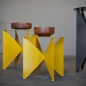 Puede incluir: Un conjunto de mesas auxiliares modernas. Dos mesas son de color amarillo brillante con un dise&ntilde;o geom&eacute;trico, cada una con un cuenco de madera. Una tercera mesa es negra con un dise&ntilde;o geom&eacute;trico similar. Una silla negra en el fondo.