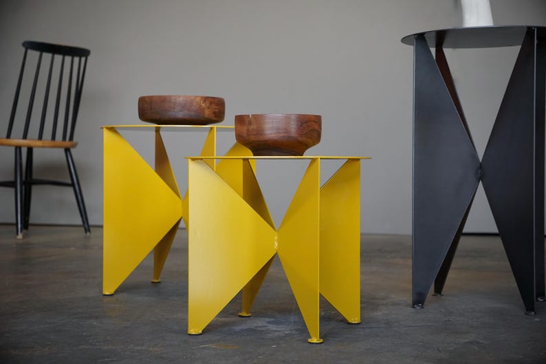 Puede incluir: Un conjunto de mesas auxiliares modernas. Dos mesas son de color amarillo brillante con un dise&ntilde;o geom&eacute;trico, cada una con un cuenco de madera. Una tercera mesa es negra con un dise&ntilde;o geom&eacute;trico similar. Una silla negra en el fondo.