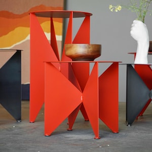 Puede incluir: Una colección de mesas auxiliares modernas de metal en rojo y negro. Las mesas tienen diseños geométricos con patas triangulares y tableros circulares. Una mesa sostiene un cuenco de madera, y otra tiene un jarrón blanco en forma de mano.