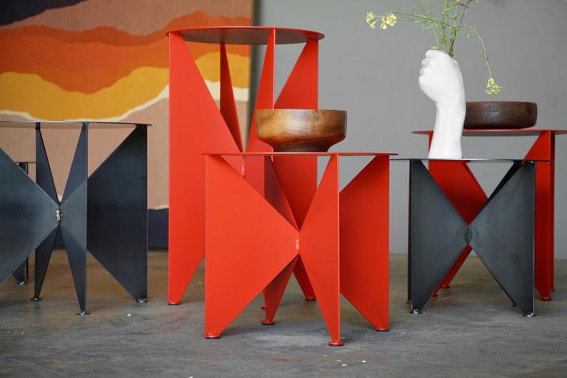 Puede incluir: Una colecci&oacute;n de mesas auxiliares modernas de metal en rojo y negro. Las mesas tienen dise&ntilde;os geom&eacute;tricos con patas triangulares y tableros circulares. Una mesa sostiene un cuenco de madera, y otra tiene un jarr&oacute;n blanco en forma de mano.