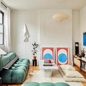 May include: A modern living room with a teal tufted sofa, a marble coffee table, and two framed abstract art prints. The room features a white ceiling light, a floor lamp, and a wooden cabinet with books and decor. The walls are white, and the floor is hardwood.