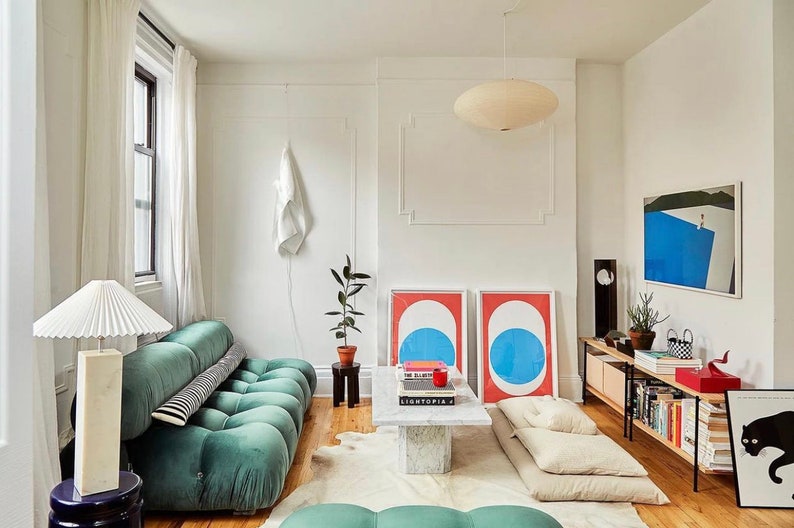 May include: A modern living room with a teal tufted sofa, a marble coffee table, and two framed abstract art prints. The room features a white ceiling light, a floor lamp, and a wooden cabinet with books and decor. The walls are white, and the floor is hardwood.