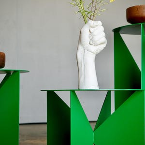 Puede incluir: Mesas auxiliares geom&eacute;tricas de color verde brillante con tapas redondas. Un jarr&oacute;n blanco en forma de mano sostiene flores amarillas. Dos cuencos de madera est&aacute;n sobre las mesas. Dise&ntilde;o moderno y minimalista.