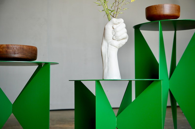 Puede incluir: Mesas auxiliares geom&eacute;tricas de color verde brillante con tapas redondas. Un jarr&oacute;n blanco en forma de mano sostiene flores amarillas. Dos cuencos de madera est&aacute;n sobre las mesas. Dise&ntilde;o moderno y minimalista.