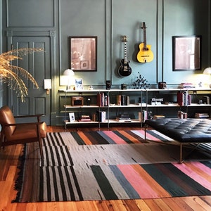 May include: A room with a teal wall, hardwood floor, and a colorful striped rug. Two guitars hang on the wall above a bookshelf filled with books and decorative items. A leather chair and a black leather bench are also in the room.