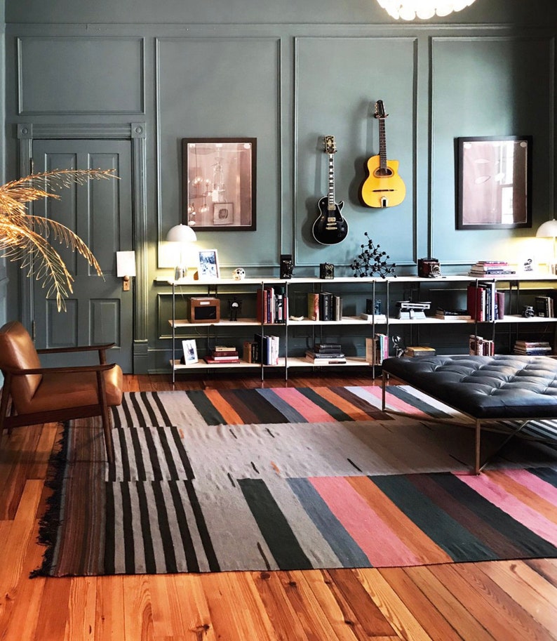 May include: A room with a teal wall, hardwood floor, and a colorful striped rug. Two guitars hang on the wall above a bookshelf filled with books and decorative items. A leather chair and a black leather bench are also in the room.