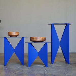 Puede incluir: Tres mesas auxiliares azules modernas con dise&ntilde;os geom&eacute;tricos. Cada mesa tiene una parte superior plana y patas angulares. Dos mesas tienen un cuenco de madera encima. Las mesas son de diferentes alturas y tama&ntilde;os.