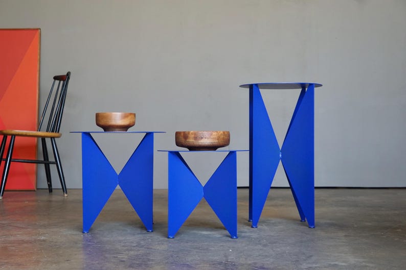 Puede incluir: Tres mesas auxiliares azules modernas con dise&ntilde;os geom&eacute;tricos. Cada mesa tiene una parte superior plana y patas angulares. Dos mesas tienen un cuenco de madera encima. Las mesas son de diferentes alturas y tama&ntilde;os.