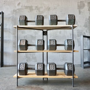 May include: A weightlifting rack with three shelves, each holding two dumbbells. The dumbbells are dark gray and marked with their weight: 80 lbs, 90 lbs, and 100 lbs. The shelves are made of light-colored wood, and the frame is black metal.