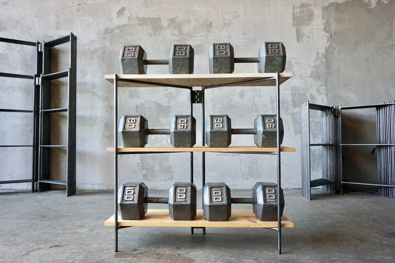 May include: A weightlifting rack with three shelves, each holding two dumbbells. The dumbbells are dark gray and marked with their weight: 80 lbs, 90 lbs, and 100 lbs. The shelves are made of light-colored wood, and the frame is black metal.