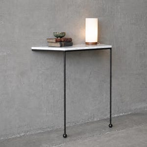 May include: A modern, minimalist side table with a white marble top and black metal legs. The table holds a small lamp, books, and a decorative bowl. The lamp has a cylindrical white shade and a wooden base. The table is against a gray wall.