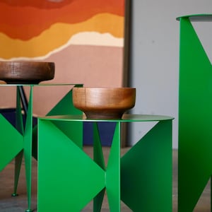 Puede incluir: Tres mesas auxiliares modernas de metal verde con dise&ntilde;os geom&eacute;tricos. Cada mesa tiene una tapa circular y un cuenco de madera. El fondo presenta una pintura abstracta con tonos naranja y marr&oacute;n.