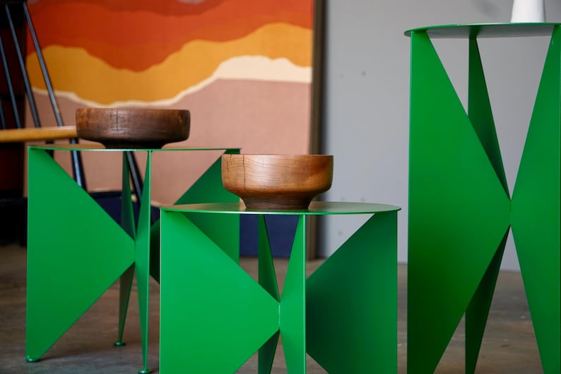 Puede incluir: Tres mesas auxiliares modernas de metal verde con dise&ntilde;os geom&eacute;tricos. Cada mesa tiene una tapa circular y un cuenco de madera. El fondo presenta una pintura abstracta con tonos naranja y marr&oacute;n.