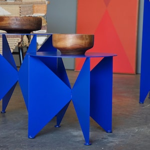 Puede incluir: Dos mesas auxiliares modernas de color azul vibrante con un llamativo dise&ntilde;o geom&eacute;trico. Cada mesa tiene una tapa circular y una base de metal plegado. Cuencos de madera sobre las mesas. Un conjunto decorativo.
