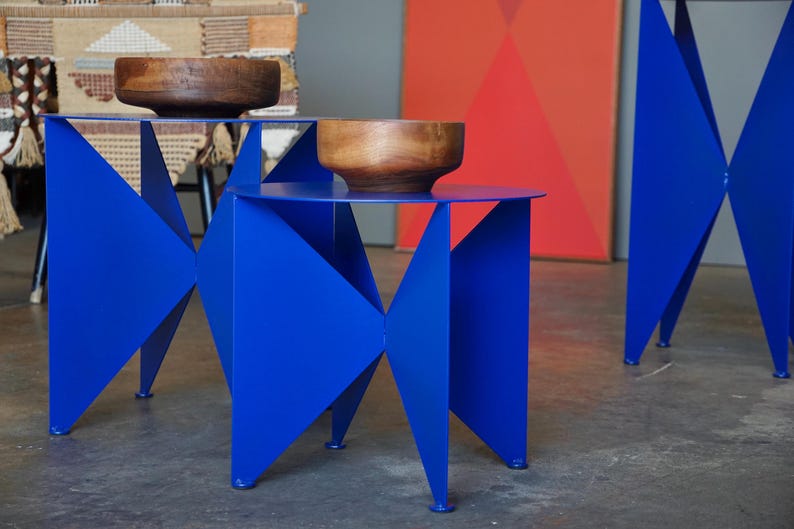 Puede incluir: Dos mesas auxiliares modernas de color azul vibrante con un llamativo dise&ntilde;o geom&eacute;trico. Cada mesa tiene una tapa circular y una base de metal plegado. Cuencos de madera sobre las mesas. Un conjunto decorativo.
