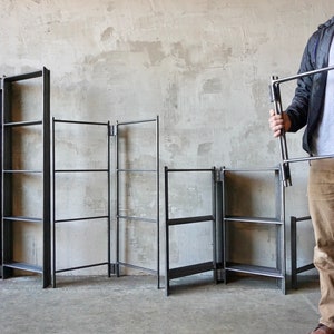 May include: Several black metal shelving units are displayed against a neutral-toned wall. One unit is held by a person. The shelves have a minimalist design, with a rectangular frame and horizontal supports. The units vary in size and configuration.