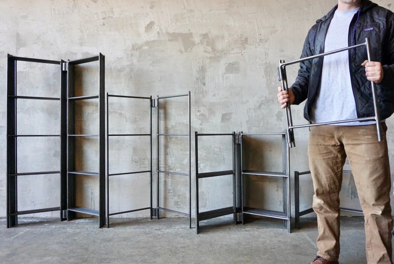 May include: Several black metal shelving units are displayed against a neutral-toned wall. One unit is held by a person. The shelves have a minimalist design, with a rectangular frame and horizontal supports. The units vary in size and configuration.