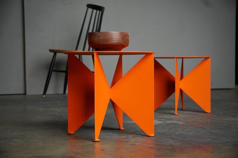 Puede incluir: Dos mesas auxiliares geom&eacute;tricas de color naranja brillante con un cuenco de madera encima. Las mesas tienen un dise&ntilde;o angular &uacute;nico y son de metal. Una silla negra est&aacute; en el fondo.