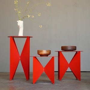 Puede incluir: Tres mesas auxiliares de metal rojo con un dise&ntilde;o geom&eacute;trico &uacute;nico. Las mesas tienen tapas redondas y patas angulares. Una mesa sostiene un jarr&oacute;n blanco con flores, y las otras sostienen cuencos de madera. Las mesas est&aacute;n sobre un suelo gris.