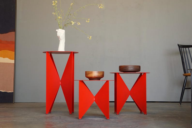 Puede incluir: Tres mesas auxiliares de metal rojo con un dise&ntilde;o geom&eacute;trico &uacute;nico. Las mesas tienen tapas redondas y patas angulares. Una mesa sostiene un jarr&oacute;n blanco con flores, y las otras sostienen cuencos de madera. Las mesas est&aacute;n sobre un suelo gris.