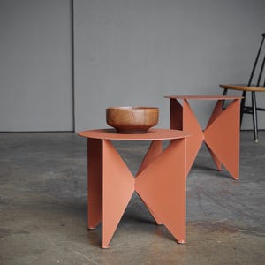 Puede incluir: Dos mesas auxiliares de color coral con dise&ntilde;os geom&eacute;tricos. Una mesa tiene un cuenco de madera encima. Una silla negra con asiento de madera est&aacute; en el fondo. Las mesas son de metal.