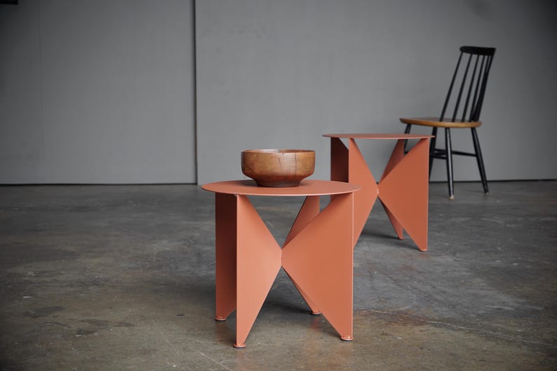 Puede incluir: Dos mesas auxiliares de color coral con dise&ntilde;os geom&eacute;tricos. Una mesa tiene un cuenco de madera encima. Una silla negra con asiento de madera est&aacute; en el fondo. Las mesas son de metal.