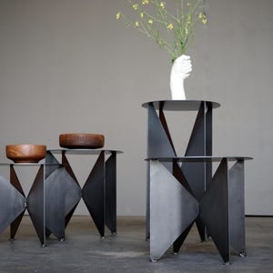 Puede incluir: Tres mesas auxiliares geom&eacute;tricas de metal con acabado oscuro. Dos cuencos de madera sobre una mesa. Un jarr&oacute;n blanco con forma de mano sostiene flores amarillas sobre otra mesa. Una silla negra en el fondo.