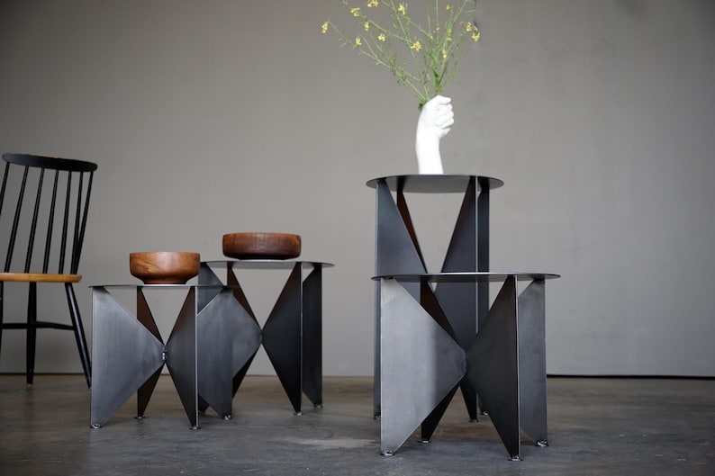 Puede incluir: Tres mesas auxiliares geom&eacute;tricas de metal con acabado oscuro. Dos cuencos de madera sobre una mesa. Un jarr&oacute;n blanco con forma de mano sostiene flores amarillas sobre otra mesa. Una silla negra en el fondo.