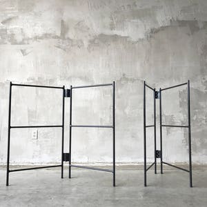 May include: A black metal folding screen with a minimalist design. The screen has a series of rectangular frames connected by hinges, allowing it to be folded. The structure is set against a textured gray wall.