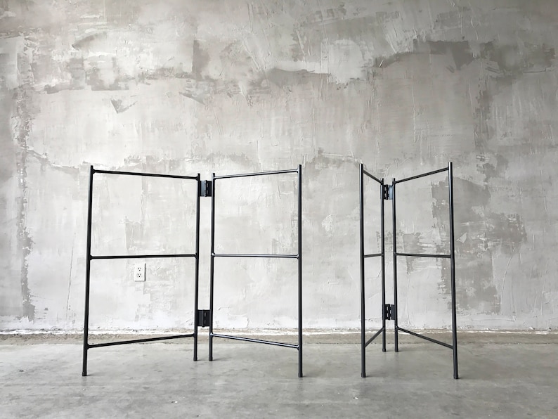 May include: A black metal folding screen with a minimalist design. The screen has a series of rectangular frames connected by hinges, allowing it to be folded. The structure is set against a textured gray wall.