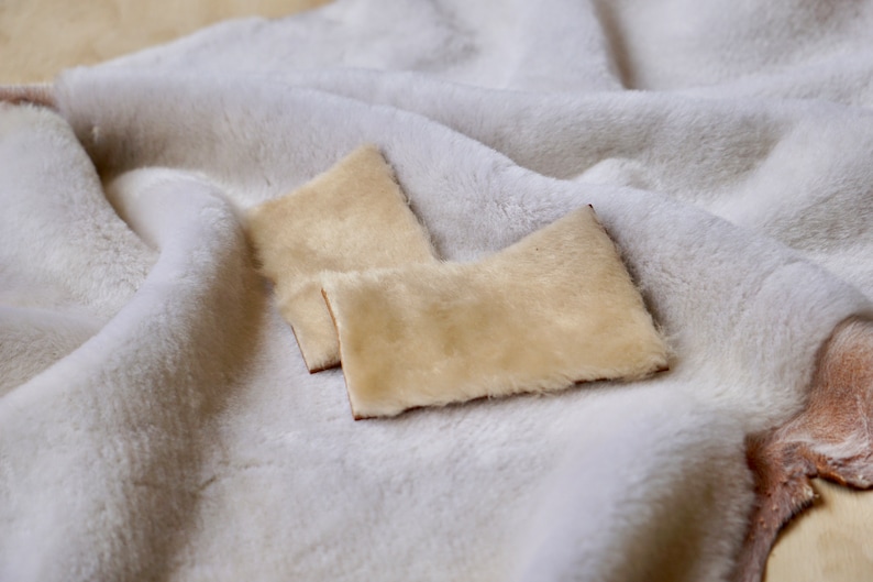 May include: Two small, cream-colored, fluffy fur squares on a white fluffy surface.
