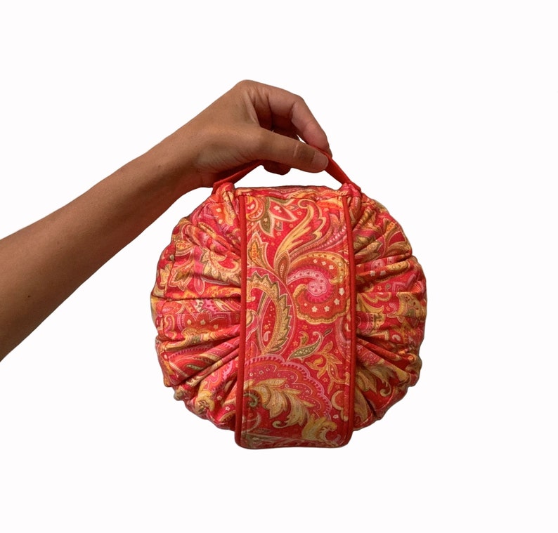 May include: A red and gold paisley patterned round cushion with a fabric handle. The cushion is made of a soft, plush fabric and has a quilted design.