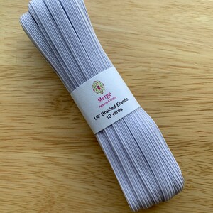May include: A bundle of braided elastic in a light lavender color, wrapped with a white label. The label reads "Merge Patterns & Crafts" and "1/4" Braided Elastic 10 yards". The elastic is on a light brown wooden surface.