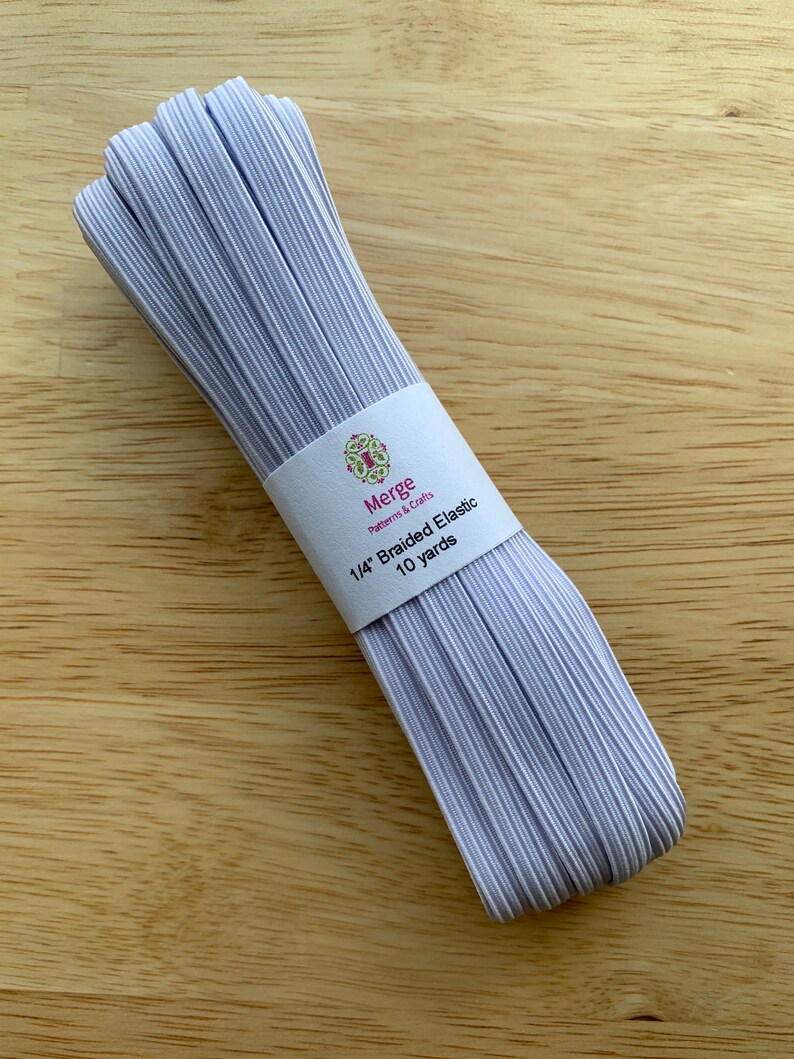 May include: A bundle of braided elastic in a light lavender color, wrapped with a white label. The label reads "Merge Patterns & Crafts" and "1/4" Braided Elastic 10 yards". The elastic is on a light brown wooden surface.